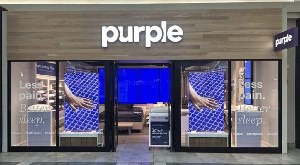 Purple Mattress retail store storefront with brand signage and display showcasing Purple Mattress products with tagline 'Less pain. Better sleep.' and promotional offers